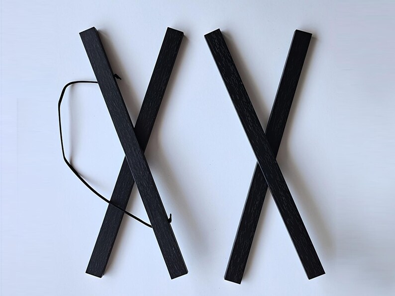 Pu&ograve; includere: Due portaposter in legno nero, ciascuno con un design a croce, sono mostrati su uno sfondo bianco. I portaposter sono collegati da uno spago nero.