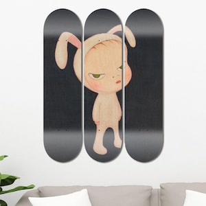 Op de afbeelding: Drie zwarte skateboard decks met een afbeelding van een kind in een roze konijnenkostuum. Het kind heeft een serieuze uitdrukking en kijkt naar links.