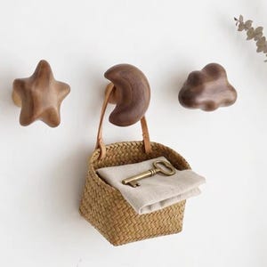 May include: Three dark brown wooden wall hooks shaped like a star, a crescent moon, and a cloud. A small woven straw basket with a light tan leather handle hangs from the moon hook, containing a folded beige cloth and a brass-colored metal key.