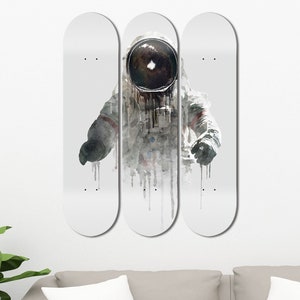 May include: Three white skateboard decks with a watercolor illustration of an astronaut in a spacesuit. The astronaut is wearing a helmet with a black visor. The illustration is in shades of gray and white.