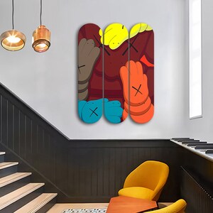 Op de afbeelding: Drie skateboard decks met kleurrijke abstracte kunst. De kunst is in een kleurenschema van rood, geel, blauw en bruin en toont een cartoonachtig personage met een groot hoofd en handen.