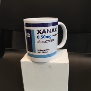 Può includere: Una tazza in ceramica bianca con un design blu e bianco che dice "Xanax 0,50mg compresse alprazolam 20 Compresse USO ORALE" e un logo Pfizer.