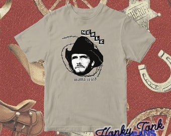 May include: A light gray t-shirt with a black and white image of a man wearing a cowboy hat. The text "MERLE" is above the image and "mama tried" is below the image.