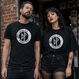 May include: Two black short-sleeved t-shirts featuring a circular white graphic on the chest. The graphic contains the text DOLLY PARTON in a circular border with a stylised DP monogram in the centre. The design has a distressed, worn texture.
