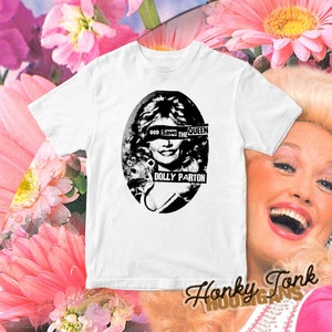 Pode incluir: Camiseta branca com uma imagem em preto e branco de Dolly Parton. A imagem tem o texto "God Save The Queen" sobre seus olhos e "Dolly Parton" abaixo. A camiseta tem o texto "Honky Tonk Hooligans" no canto inferior direito.