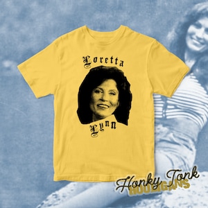 Puede incluir: Camiseta amarilla con una imagen en blanco y negro de Loretta Lynn y el texto "Loretta Lynn" en una fuente gótica.