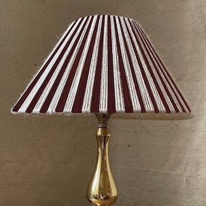 Puede incluir: Una lámpara de mesa de latón con una pantalla de lámpara a rayas blancas y burdeos. La pantalla de la lámpara está hecha de tela y tiene un diseño plisado. La lámpara está sobre una mesa de madera.