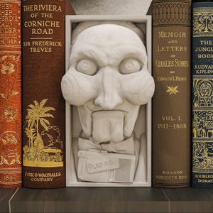 Puede incluir: Una escultura blanca de una cara de marioneta con pajarita y el texto "PLAY ME" está centrada entre libros antiguos. Los libros son de varios colores, incluyendo marrón y naranja, con letras doradas.