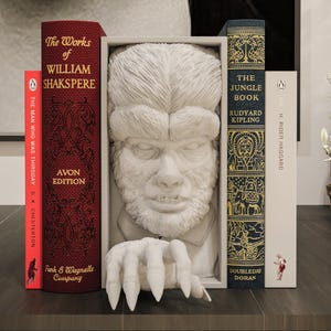 Könnte beinhalten: Ein dekoratives Bücherregal mit einer weißen Werwolf-Büste, flankiert von klassischen Büchern. Die Bücher umfassen "The Works of William Shakespeare" und "The Jungle Book". Die Bücher sind in Rot, Weiß und Blau mit goldener Schrift.