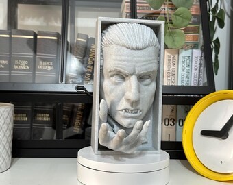May include: A gray 3D printed sculpture of a man's head and hand emerging from a rectangular frame. The sculpture is on a white circular base. The text "NUNCA TE PAR" is visible on the frame.