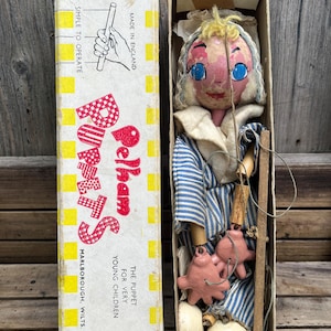 May include: A vintage Pelham Puppets marionette in its original box. The puppet has a painted face with blue eyes, yellow hair, and wears a blue and white striped outfit. The box features yellow and white checkered borders and red text.