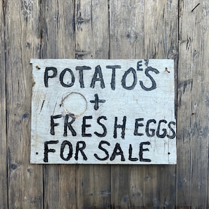 May include: A distressed wooden sign, painted with the words "POTATO'S + FRESH EGGS FOR SALE" in black. The sign is attached to a wooden door with vertical planks.