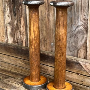 May include: Two tall, cylindrical wooden spools with a natural brown wood grain finish. Each spool features a circular metal base and a matching circular metal top, both with a dark, weathered, and slightly textured metallic appearance.