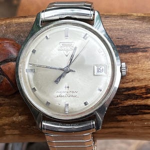 May include: A vintage Hamilton electronic wristwatch with a silver dial and a date window at the 3 o'clock position. The watch features the "ARMCO" logo and the text "HAMILTON electronic". The watch has a silver metal strap.