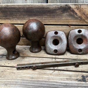 May include: A set of two spherical metal door knobs with a dark, rusted finish, accompanied by two matching square-shaped metal backplates with screw holes and a single rectangular metal spindle bar featuring two small screws for assembly.