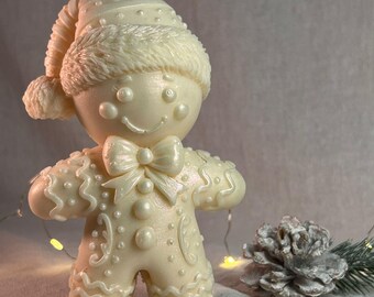 May include: A cream-colored gingerbread man candle with a Santa hat and bow tie. The candle has a smiling face and is decorated with white icing details. A pine cone and string lights are in the background.
