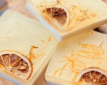 May include: Three square bars of soap with dried lemon slices and flower petals embedded in the soap. The soap is a light yellow color.