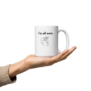 Könnte beinhalten: Eine weiße Keramiktasse mit einer schwarzen Umrandung einer Hand, die sich ans Ohr hält, und dem Text "I'm all ears."
