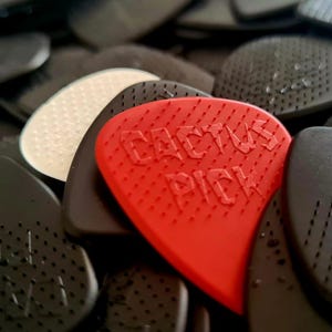 Op de afbeelding: Close-up van een verzameling gitaar plectrums. Een felrode plectrum met de tekst "CACTUS PICK" staat in het midden, omringd door vele zwarte plectrums. Sommige plectrums hebben een textuur. Voor muziekinstrumenten.