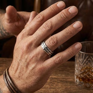 Puede incluir: Un anillo plateado con un diseño de cruz se lleva en un dedo. El anillo tiene una apariencia en capas. Una pulsera de cuero marrón está en la muñeca. Un vaso de líquido ámbar está sobre una superficie de madera.