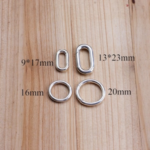 Könnte beinhalten: Vier silberfarbene Metallringe und -schlaufen in verschiedenen Größen werden auf einer Holzoberfläche gezeigt. Die Größen sind mit 9*17mm, 13*23mm, 16mm und 20mm angegeben. Es handelt sich wahrscheinlich um Schmuckzubehör oder Hardware.