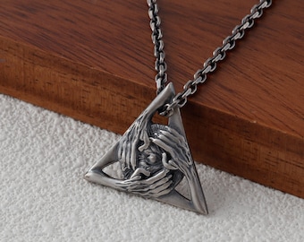 Puede incluir: Un collar con colgante plateado. El colgante triangular presenta un diseño detallado de manos que rodean una figura central. El collar tiene una cadena y se muestra sobre una superficie de madera.