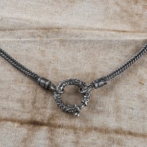 May include: Close-up of a silver necklace with a decorative circular clasp. The chain has a woven design, and the clasp features intricate scrollwork. The necklace is displayed on a neutral-toned fabric background, highlighting the jewelry's details.