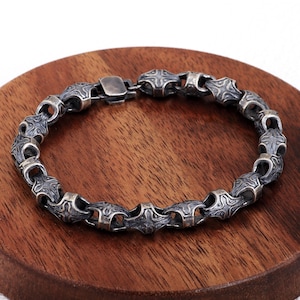 May include: A silver bracelet with a detailed, patterned design. The bracelet is composed of interconnected, stylized links. The clasp is a simple, rectangular design. The bracelet is displayed on a wooden surface.