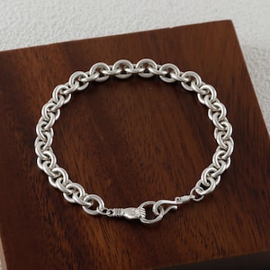 May include: A silver bracelet with a chain link design. The bracelet features a series of interconnected, rounded links. The clasp is a hook and eye closure. The bracelet is displayed on a dark wooden surface.