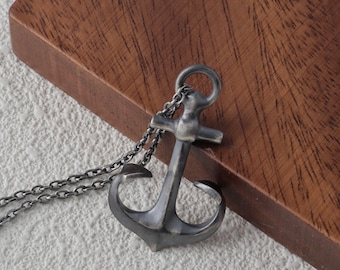 May include: A silver-coloured anchor pendant necklace. The anchor design has a classic shape with a ring at the top and a chain. The necklace is displayed on a wooden surface.
