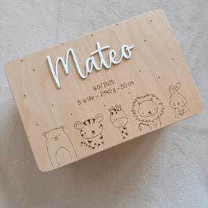 Puede incluir: Una caja de madera clara con el nombre "Mateo" en escritura blanca. La caja presenta ilustraciones de animales grabadas y la fecha "16.07.2025", peso y altura.