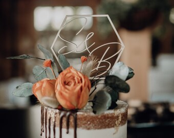 May include: A white wedding cake with chocolate ganache dripping down the sides. The cake is decorated with orange flowers and greenery. A wooden hexagon topper with the initials "S & B" is on top of the cake.