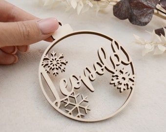 Könnte beinhalten: Ein runder Weihnachtsbaumschmuck aus Holz mit dem Namen "Leopold" in Kursivschrift. Der Schmuck ist mit Schneeflocken verziert.