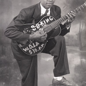 Könnte beinhalten: Schwarz-Weiß-Fotografie von B.B. King, einem Bluesmusiker, mit einer Gitarre. Die Gitarre ist schwarz mit weißer Schrift, die "B.B. KING" und "W.D.I.A 530 P.M." lautet. Er trägt einen Anzug mit Krawatte, ein Fuß ruht auf einem Hocker.