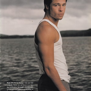 Könnte beinhalten: Ein Magazinbild zeigt einen Mann in einem weißen Tanktop und grauer Hose, der sich in der Nähe eines Gewässers aufhält. Der Mann schaut in die Kamera. Text am unteren Bildrand identifiziert Brad Pitt und die Fotografin Annie Leibovitz.