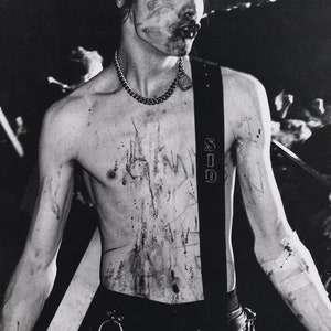 May include: A black and white photo of a man with a guitar strap and a chain necklace. He is covered in what appears to be blood or paint. The photo is taken from the chest up.