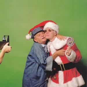 May include: Two men, one wearing a Santa Claus costume and the other wearing a blue denim jacket and a blue and white hat, are hugging. The man in the Santa costume is holding a purple and white lollipop.