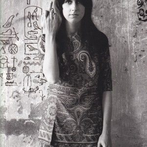 May include: A woman with long dark hair wearing a paisley patterned dress and pants stands in front of a wall with graffiti. She is giving the middle finger.