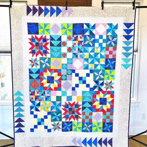 May include: A colorful patchwork quilt with a variety of geometric patterns, including stars, squares, and triangles. The quilt features a white border and is displayed on a black stand. The quilt's colors include blue, red, yellow, green, and purple.
