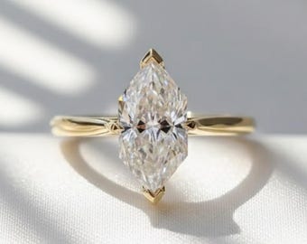 May include: A marquise-cut diamond engagement ring with a slender gold band. The diamond is set with pointed prongs at the top and bottom, and the ring is displayed on a white, textured surface.