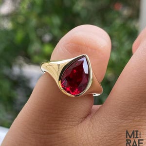 Peut inclure: Une bague en or avec une grosse pierre précieuse rouge foncé en forme de goutte d'eau. La bague a un anneau et un sertissage en or lisse et poli. La pierre précieuse est le point central, reflétant une couleur vibrante. La bague est une pièce de déclaration.
