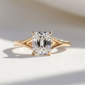 Może przedstawiać: Złoty pierścionek z dużym diamentem o prostokątnym szlifie. Przezroczysty diament osadzony jest w czteroramiennej oprawie. Opaska ma konstrukcję z rozciętym trzonkiem, dodającą nowoczesnego akcentu do klasycznego stylu pierścionka zaręczynowego.