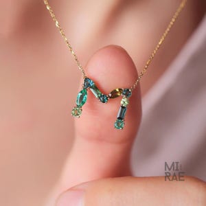 Puede incluir: Un collar dorado con la letra "M" como colgante, adornado con piedras preciosas verdes y azules. Las piedras preciosas tienen diferentes formas y tamaños, creando un diseño colorido. El collar está sostenido por el dedo de una persona.