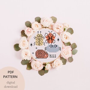 Könnte beinhalten: Ein Kreuzstichmuster mit einem Schmetterling, einer Schnecke, einer Spinne und einem Käfer. Das Muster ist in einem Reifen und von rosa Rosen umgeben.