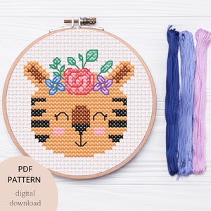 May include: A cross-stitch pattern of a cute tiger with a floral crown. The pattern is in a wooden embroidery hoop and includes floss in various colors.