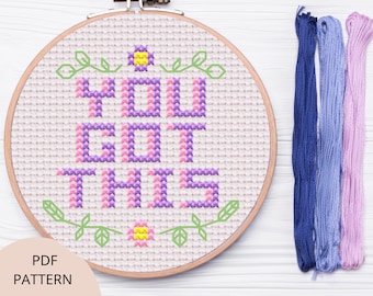 Puede incluir: Un aro de bordado de punto de cruz con la frase "You Got This" en hilo rosa y morado. El aro está sobre una superficie de madera blanca con cuatro madejas de hilo de bordar en azul, morado y rosa.