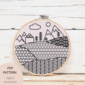 May include: A black and white geometric cross stitch pattern of a mountain landscape with a pink background. The pattern includes mountains, trees, a house, a cloud, and a sun. The pattern is in a round embroidery hoop.