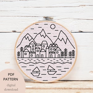 Op de afbeelding: Een zwart-witte lijntekening van een bergdorp met een meer en boten. Het ontwerp is op een roze en witte stoffen achtergrond in een ronde borduurring.