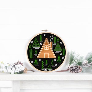 May include: A black and white embroidery hoop with a design of a cabin in a forest. The cabin is brown and the trees are green. The background is black and the design is outlined in white.