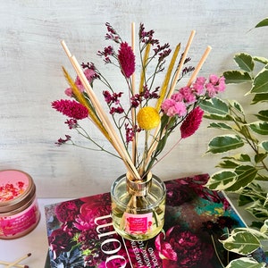 May include: A clear glass diffuser bottle with a gold lid and reeds. The bottle is filled with a light yellow liquid. The reeds are surrounded by dried flowers in shades of pink, yellow, and red. The diffuser is sitting on a book with the word "bloom" printed on the cover.